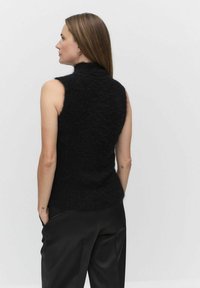 Musta hihaton neulepaita korkealla kauluksella, jossa on pehmeä, pörröinen pinta. Takana paita istuu tiiviisti vyötäröltä ja levenee hieman.