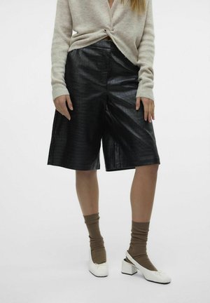Frau trägt einen beigen Strickpullover, schwarze knielange Shorts aus Kunstleder, braune Socken und weiße Slingback-Schuhe mit Blockabsatz.