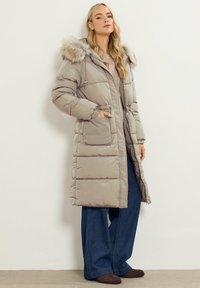 Abrigo largo acolchado beige con capucha de piel, bolsillos frontales y cierre de cremallera. Lleva puesto unos pantalones vaqueros oscuros holgados y unos zapatos marrones.