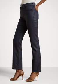 Tumši zili džinsa zvēri biksēs ar pieguļošu vidukli, piecām kabatām un zelta toņa apkalumiem; kombinētas ar brūnām augstpapēžu sandaletēm ar siksniņām.