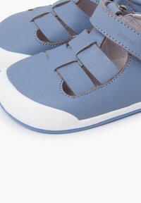 Zapatos de cuero azul con detalles recortados y una correa de Velcro; suela de goma blanca con acentos azules; textura suave y bordes cosidos.