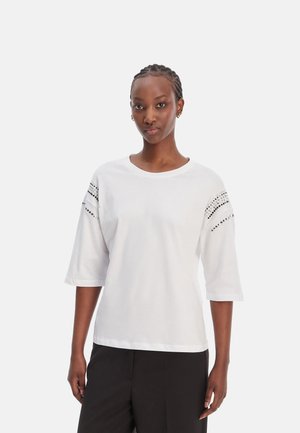 Donna con capelli intrecciati che indossa una maglia bianca con maniche a gomito e strisce borchiate nere sulle spalle e pantaloni neri su uno sfondo semplice.