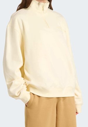 Kobieta w jasnobeżowej bluzie z długim rękawem i zamkiem do połowy wysokości oraz w camelowych spodniach, z rękami w kieszeniach, stojąca na tle gładkiego tła.