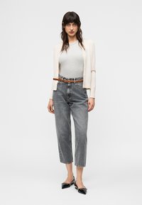 Nő törtfehér gombos kardigánban, szürke testhezálló felsőben, magasderekú, kifakult farmert barna övvel és fekete hegyes orrú lapos cipővel, fehér háttér előtt állva.