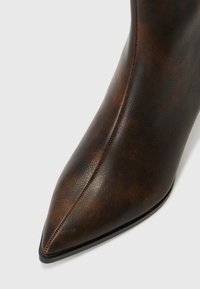 Botte en cuir marron foncé à bout pointu avec un détail de couture centrale et une semelle noire sur un fond gris clair.