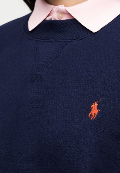 Μπλε ναυτικού πληρώματος πουλόβερ με μικρό πορτοκαλί λογότυπο παίκτη πόλο πάνω από ροζ ανοιχτό πουκάμισο με γιακά.