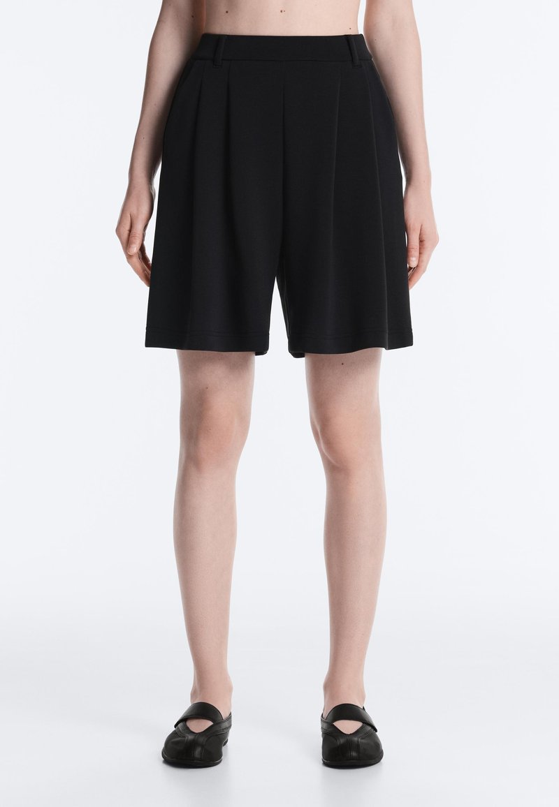 Personne portant un short plissé noir arrivant aux genoux et des chaussures noires sans lacets, debout devant un fond blanc uni.