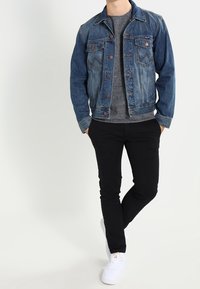 Donkerblauwe spijkerjas met twee borstzakken, gecombineerd met een grijze trui en zwarte slim-fit broek. Witte sneakers maken de look compleet.