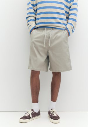 Person trägt beige Shorts mit Kordelzug, blau-weiß gestreiftes Langarmshirt, weiße Socken und braune Turnschuhe und steht mit den Händen in den Taschen.
