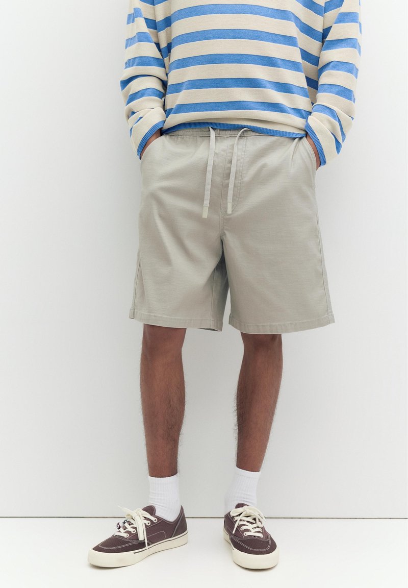 Personne portant un short beige à cordon de serrage, une chemise à manches longues rayée bleu et blanc, des chaussettes blanches et des baskets marron, debout les mains dans les poches.
