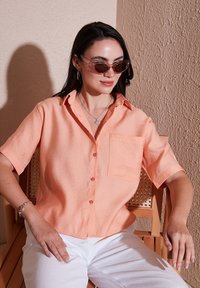Short-sleeve button-up shirt in light coral, with a chest pocket and textured fabric. Paired with white pants, on a wooden chair against a textured wall.