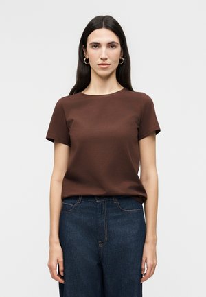 Γυναίκα με μακριά σκούρα μαλλιά που φορά χρυσούς κρίκους στα αυτιά, καφέ κοντομάνικο πουκάμισο και σκούρο μπλε τζιν, στέκεται μπροστά από ένα απλό φωτεινό φόντο.