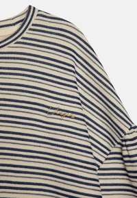 Pull rayé en crème et bleu marine, avec un motif de tricot texturé et un logo brodé en or sur le côté gauche.