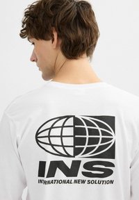 Camiseta de manga larga blanca con un gran gráfico negro de un globo y el texto "INS SOLUCIÓN NUEVA INTERNACIONAL" en la parte posterior.