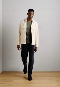 Lichtbeige jas met twee borstzakken, gedragen over een grijs shirt en donkere spijkerbroek. Model draagt donkere schoenen en staat op een houten vloer.