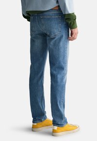 Lichtblauwe denim jeans met een rechte pasvorm, voorzien van achterzakken en een leren detail. Gecombineerd met gele sneakers op een witte achtergrond.