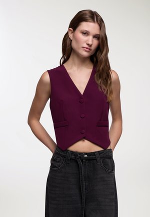 Gilet in tessuto bordeaux scuro, caratterizzato da un colletto a V, tre bottoni e due tasche frontali; design corto abbinato a jeans neri a vita alta.