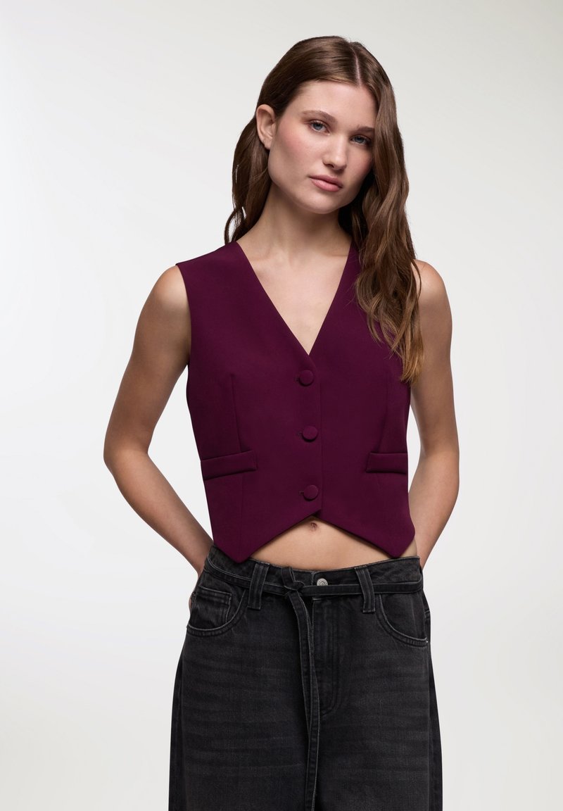 Gilet in tessuto bordeaux scuro, caratterizzato da un colletto a V, tre bottoni e due tasche frontali; design corto abbinato a jeans neri a vita alta.