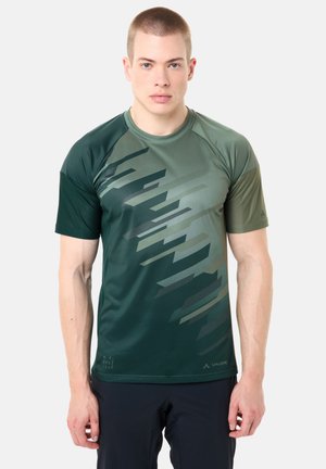 Junger Mann mit kurzen Haaren, der ein grünes, gemustertes, kurzärmliges Sportshirt und dunkle Hosen trägt, vor einem einfarbigen Hintergrund steht.