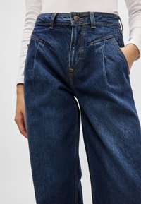 Jeans de ganga azul de cintura alta com um corte solto, apresentando pregas frontais, costura laranja e bolsos laterais. Textura suave e lavagem escura.