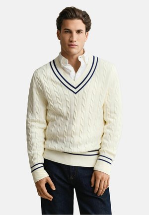 Mann trägt einen weißen Kabelstrick-V-Ausschnitt-Pullover mit marineblauen Streifen über einem weißen Hemd mit Knöpfen, kombiniert mit dunkelblauen Jeans.