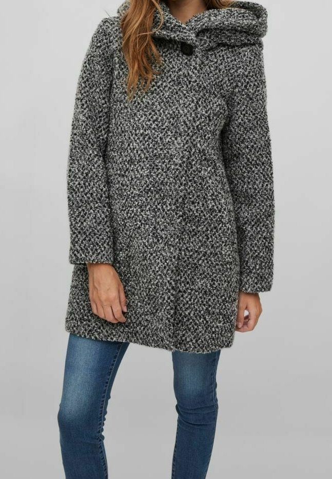 Femme portant un manteau à capuche gris texturé avec un bouton noir au col et un jean skinny bleu, debout devant un fond uni.