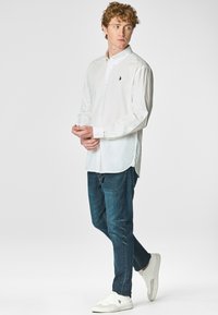 Camicetta bianca con colletto a punta, con un piccolo logo blu navy sul petto. Indossata con jeans di denim scuro e sneakers bianche.