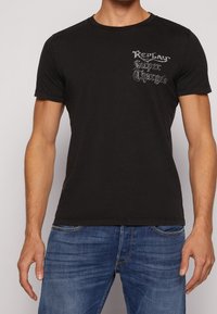 Czarna bawełniana koszulka z okrągłym dekoltem, krótkimi rękawami i nadrukowanym logo w srebrnym kolorze na piersi. Połączona z niebieskimi dżinsami.