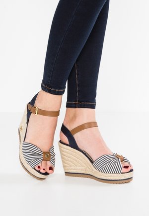 Fødder iført stribede kilehælssandaler med brune ankelremme og mørke smalle jeans mod en hvid baggrund.