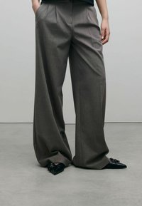 Pantalons gris à jambes larges avec des plis à la taille, fabriqués à partir d'un tissu lisse. Portés avec des chaussures à talons aiguilles noires présentant des détails froncés.