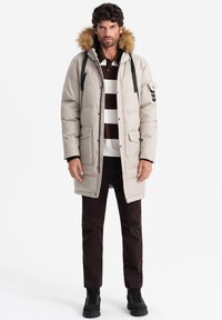 Cappotto imbottito beige chiaro con cappuccio foderato in pelliccia, dettagli neri, chiusura con zip e molteplici tasche, indossato sopra una polo a righe marroni e pantaloni scuri.