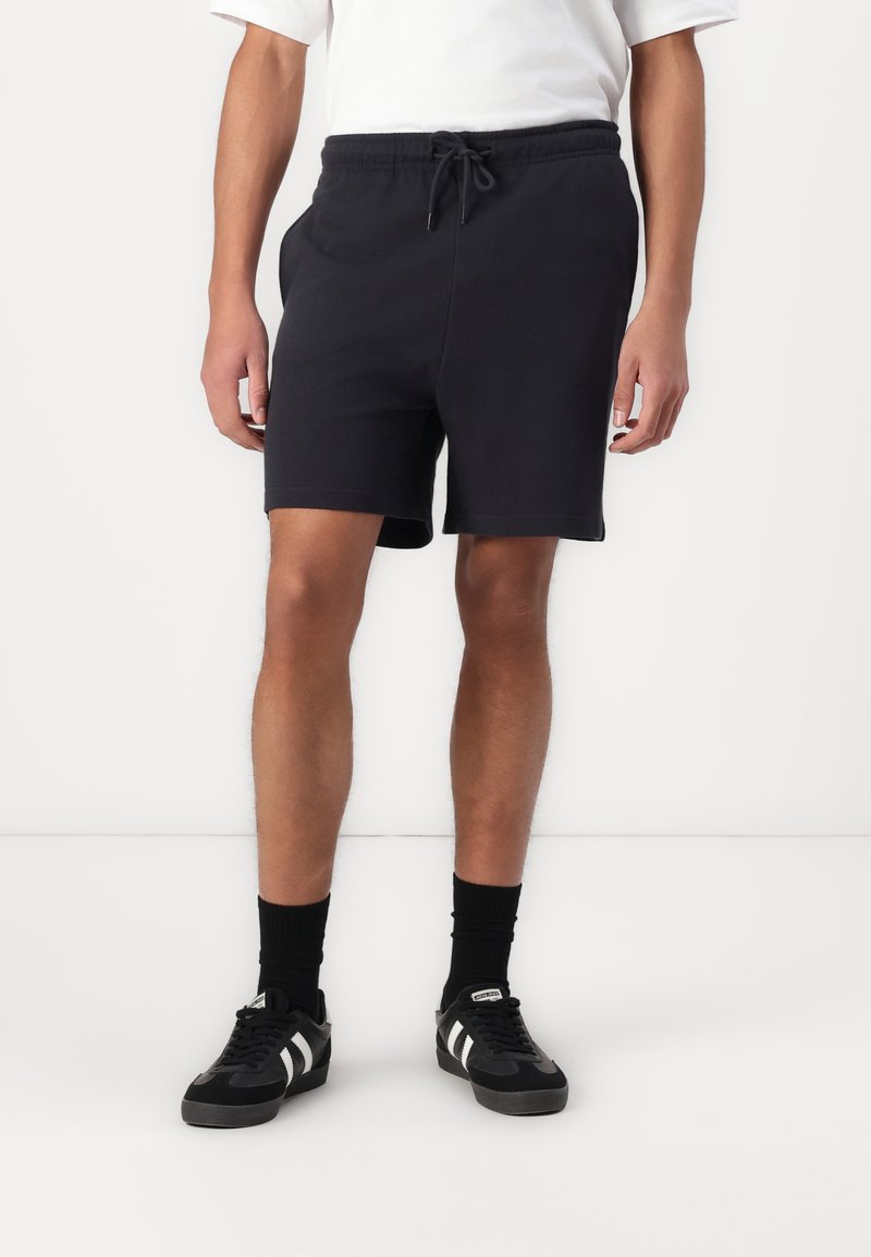 Black cotton shorts with an elastic waistband and drawstring, featuring side pockets. Paired with black sneakers and crew-length black socks.