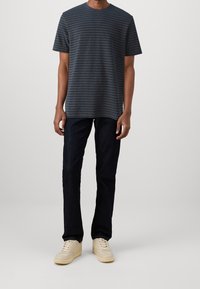 Homme portant un t-shirt rayé bleu marine et gris, un jean foncé et des baskets beiges, se tenant devant un fond clair uni.