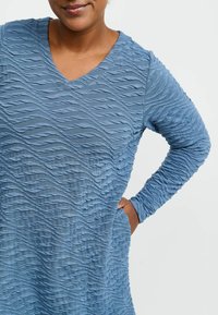 Långärmad klänning med V-ringning i texturerat blått tyg med vågiga mönster. Har en avslappnad passform och sidofickor.