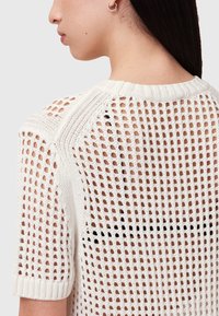 Løst strikket sweater i hvid, med korte ærmer, en ribbet krave og et mønstret ryg. Materialet virker let og åndbart.
