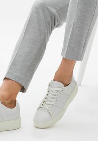Białe skórzane sneakersy o gładkiej fakturze, z okrągłym noskiem, sznurowanym zapięciem i teksturowaną białą podeszwą, zestawione z szarymi spodniami.