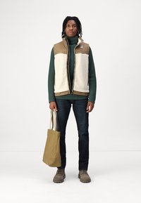Gilet in fleece marrone e crema su un maglione a coste verde, jeans scuri e scarpe grigie. Tieni una borsa a tracolla beige. Design semplice e informale.