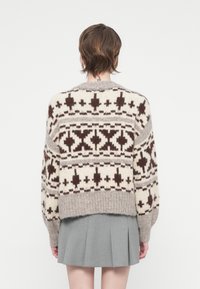 Pull en tricot crème et marron, avec des motifs géométriques, un col rond et des manches amples. Présenté avec une jupe plissée grise.