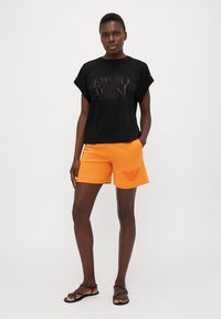 Black short-sleeve t-shirt with Emporio Armani logo. Orange shorts with textured logo detail. Model wears black sandals.