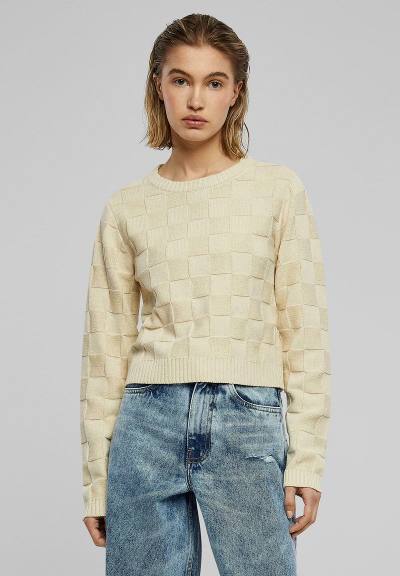Beige gebreide trui met een gestructureerd ruitpatroon, geribbelde ronde hals en lange mouwen, gecombineerd met lichtblauwe versleten jeans.