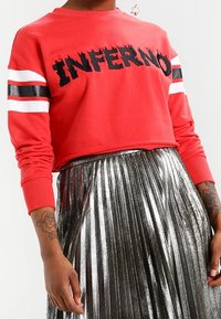 Sudadera roja corta con el texto negro "INFERNO", con mangas a rayas blancas y negras. Combinada con una falda plisada plateada metálica.