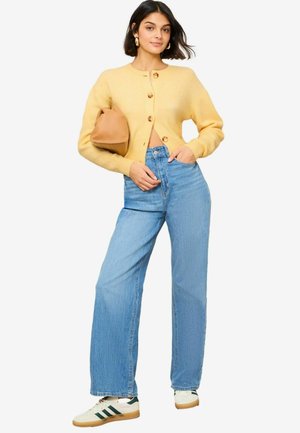 Yellow wool cardigan with round neck and wooden buttons, paired with light blue high-waisted wide-leg jeans and white sneakers with green accents.