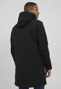 Abrigo largo negro con capucha, hecho de un tejido suave y mate. Presenta un corte recto y un diseño minimalista, sin herrajes visibles ni patrones.