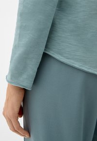 Hellblaues Langarmoberteil mit weicher Textur, abgerundetem Saum und genähten Details, kombiniert mit einem passenden blauen Stoff darunter.