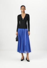 Schwarzes Langarmoberteil mit gedrehtem Vorderteil und silbernem Akzent; blauer Tüllrock; schwarze Absätze; schwarze Handtasche.