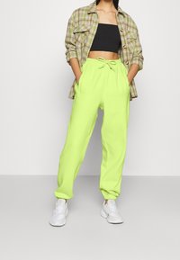 Kvinna i neongröna sweatpants med dragsnöre, svart tube top, rutigt skjorta, händerna i fickorna, och vita sneakers på en enfärgad bakgrund.