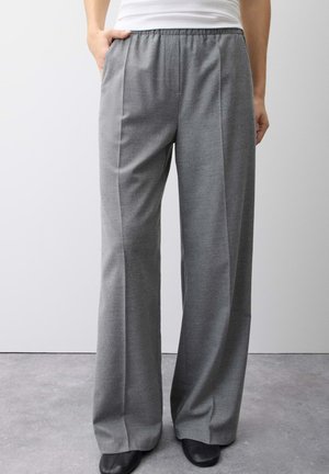 Personne portant un pantalon gris ample avec une taille élastique et des chaussures noires, debout sur un sol gris contre un mur blanc.