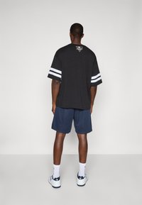 Schwarzes Kurzarmshirt mit weißen Streifen an den Ärmeln, Logo auf dem Rücken, kombiniert mit navyblauen Mesh-Shorts und weißen Sneakers mit navyblauen Akzenten.