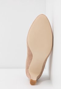 Beige suède hoge hak schoen met een spitse neus, voorzien van een gladde rubberen zool en een subtiel getextureerd grippatroon.
