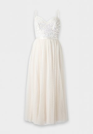 Femme portant une robe midi blanche sans manches avec un corsage à sequins et une jupe transparente à pois, debout avec des sandales à talons beiges.